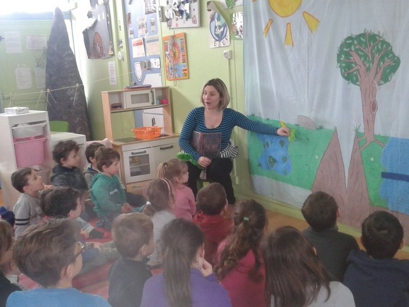 Mdiatrice scientifique racontant un conte  des enfants  l'aide d'un grand dessin accroch au tableau, les enfants sont assis par terre autour d'elle dans la classe